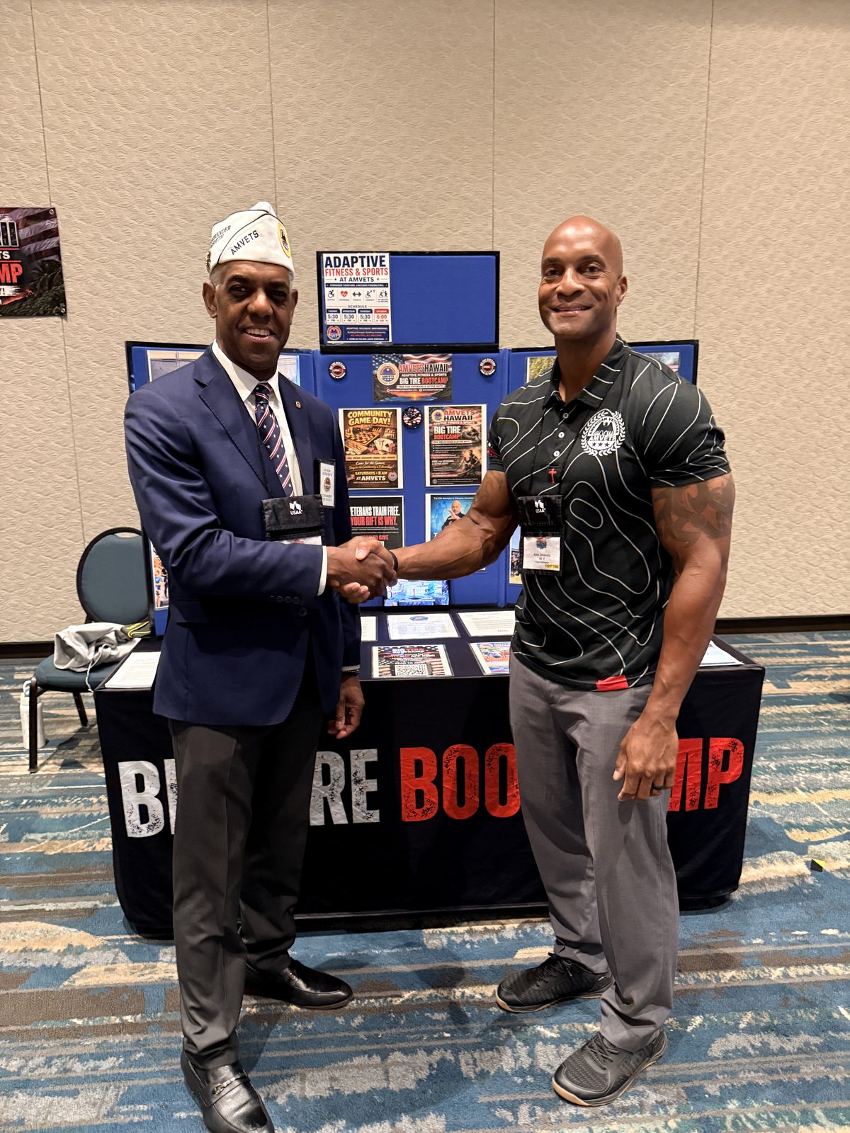 An AMVETS officer and Coach Alim shaking hands in front of the Big Tire Bootcamp exhibit.