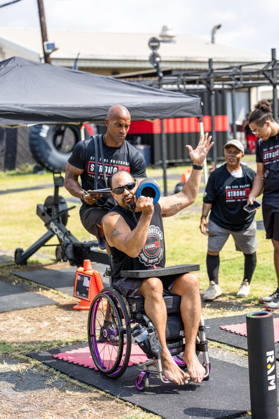 Garrett, a retired U.S. Air Force Master Sergeant, pressing a weight plate from his wheelchair while a Big Tire Bootcamp coach spots him on Warrior Field.